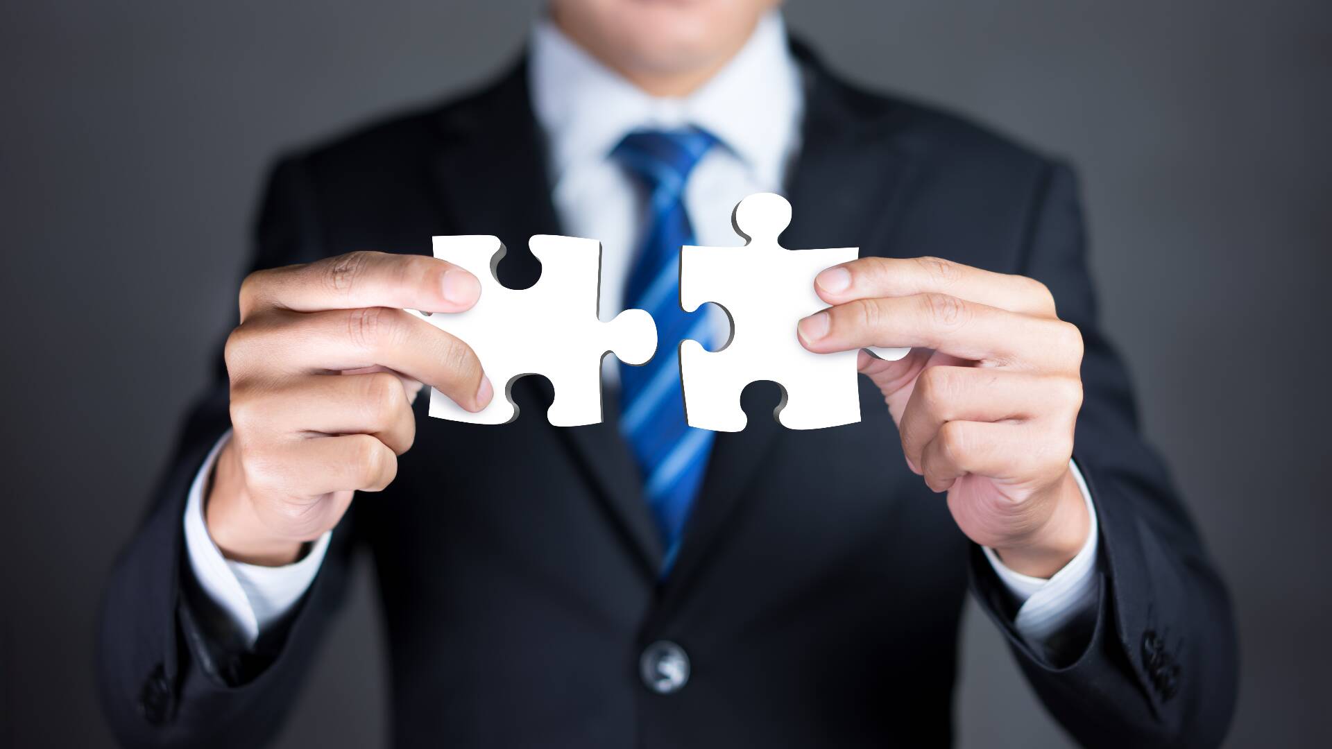 Executive in suit and blue tie holds connecting puzzle pieces representing the merging of two companies or a joint venture or partnership.